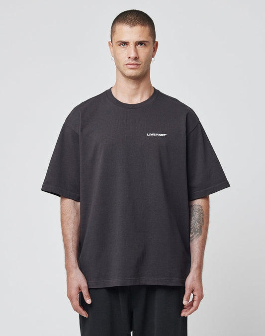 A man with short hair faces forward, wearing the LIVE FAST DIE YOUNG LFDY Cinema Tee—an oversize black cotton T-shirt with LIVE FAST in white on the left chest. A tattoo is visible on his left forearm against a plain light gray background.