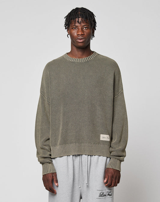 A man with short twists wears the LIVE FAST DIE YOUNG LFDY Knit Sweater, an oversized ribbed olive-green piece in 100% cotton, paired with light gray sweatpants. He stands facing forward against a plain light gray background. | model