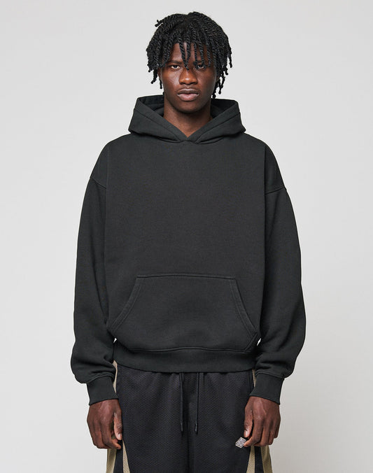 A person with short, twisted hair faces forward, wearing a black Basic Blank Hooded by LIVE FAST DIE YOUNG and black shorts with beige stripes and a flag label, set against a plain light gray background. | model