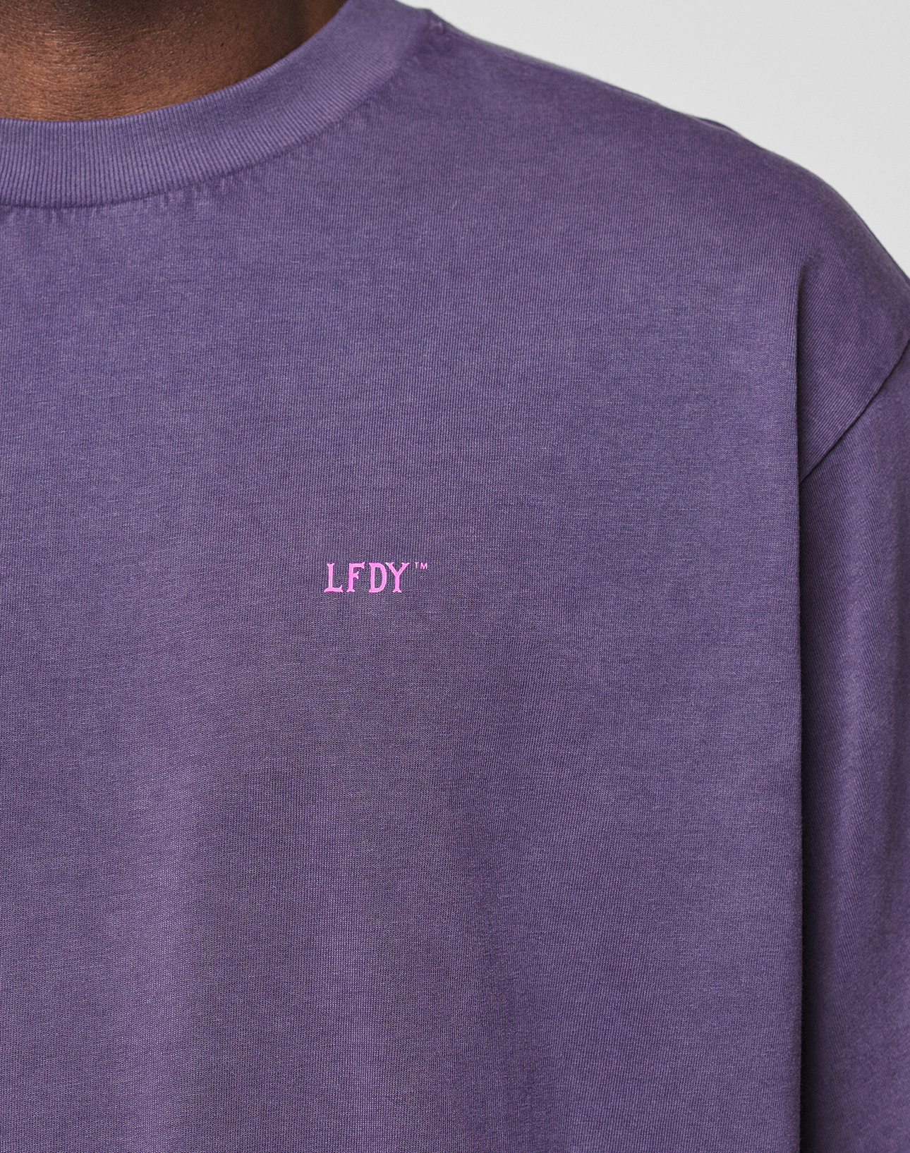 A close-up shows a person wearing the Basic Evidence Tee by LIVE FAST DIE YOUNG. The neck, shoulder, and upper torso are visible against a plain background. The tee is purple with small pink LFDY™ text on the chest.