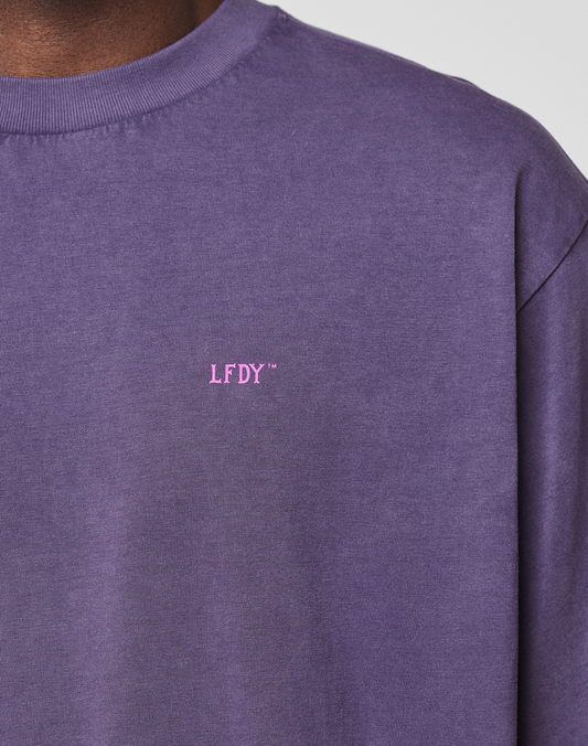 A close-up shows a person wearing the Basic Evidence Tee by LIVE FAST DIE YOUNG. The neck, shoulder, and upper torso are visible against a plain background. The tee is purple with small pink LFDY™ text on the chest.