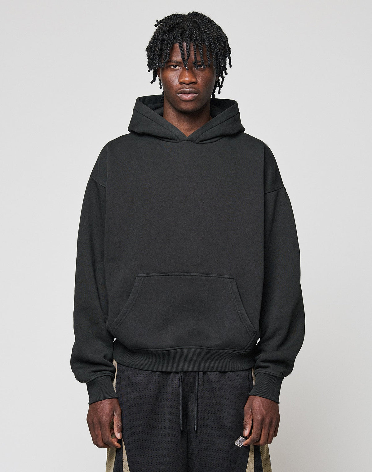 A person with short, twisted hair faces forward, wearing a black Basic Blank Hooded by LIVE FAST DIE YOUNG and black shorts with beige stripes and a flag label, set against a plain light gray background. | model