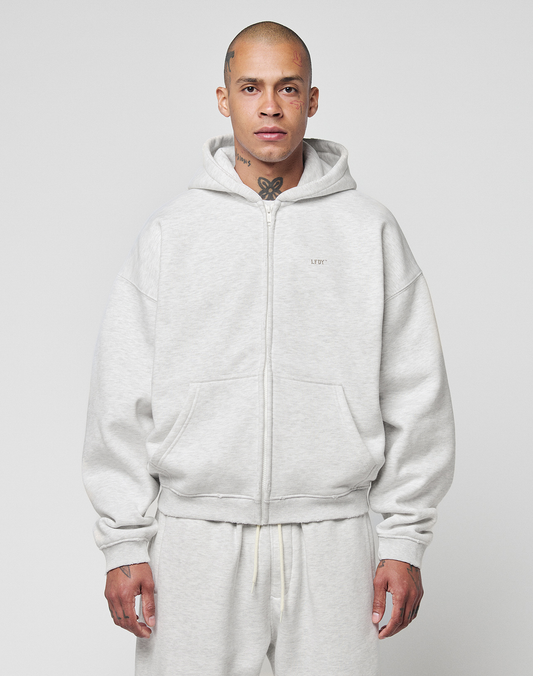 A person with a shaved head and visible tattoos models the LIVE FAST DIE YOUNG Basic Hooded Zip in light gray, paired with matching sweatpants, standing against a plain background and looking directly at the camera with a neutral expression. | model