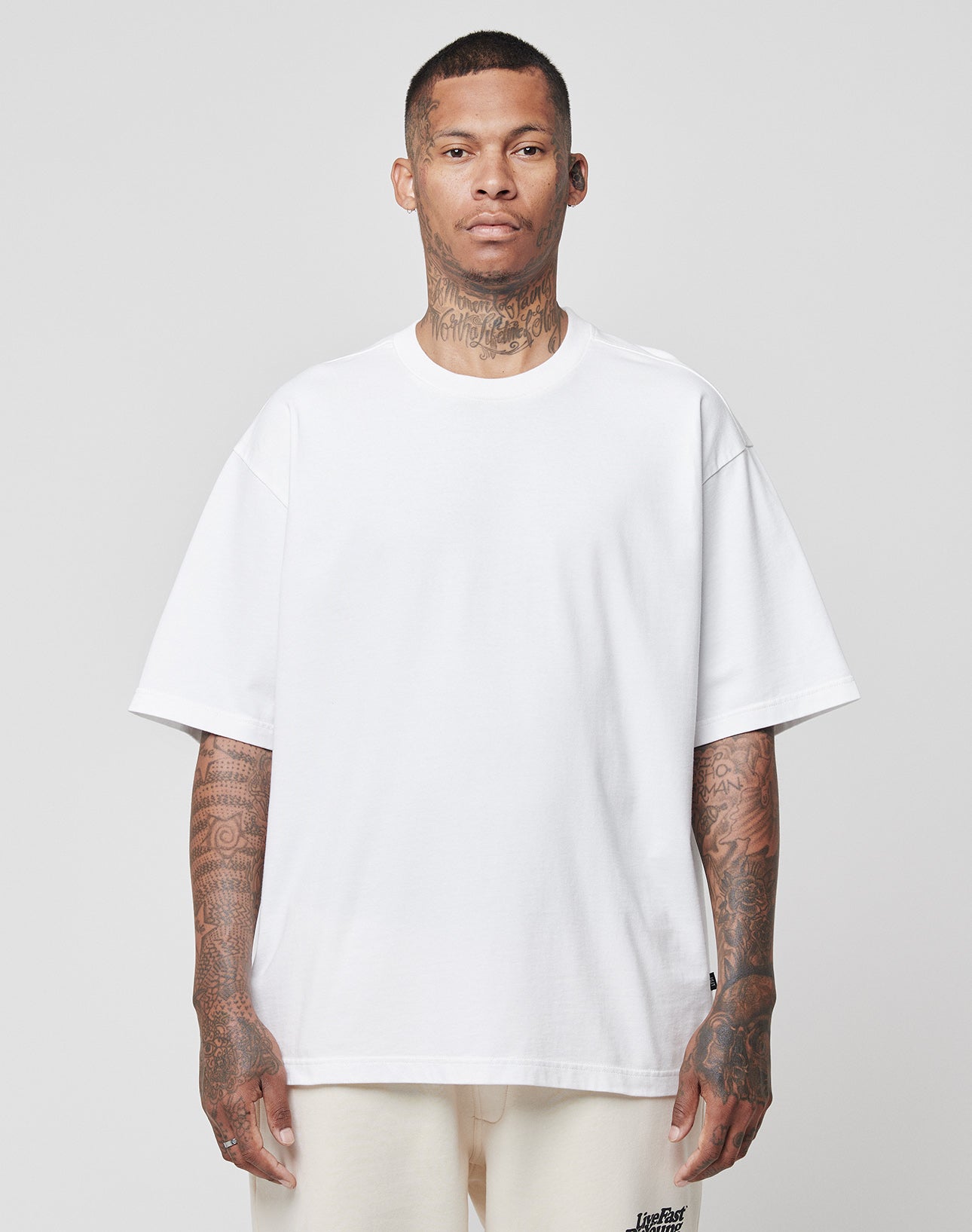 A man with short hair and visible neck and arm tattoos faces forward in an oversized LIVE FAST DIE YOUNG Basic Heavy Tee (100% Baumwolle, plain white) and off-white pants, standing against a plain light gray background.