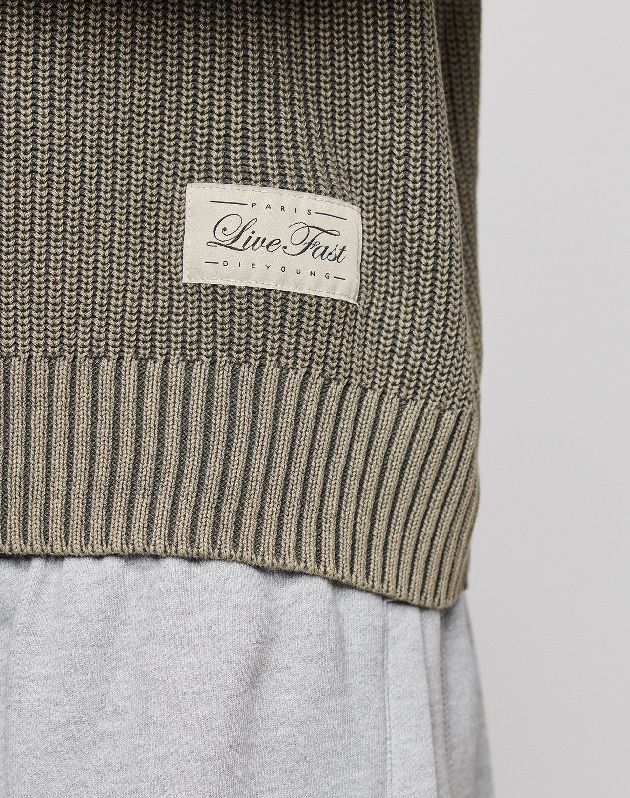 Close-up of the LFDY Knit Sweater by LIVE FAST DIE YOUNG, featuring an oversize fit, dropped shoulders, and a beige patch with "Live Fast" and "Paris DeYoung," styled over light gray pants.
