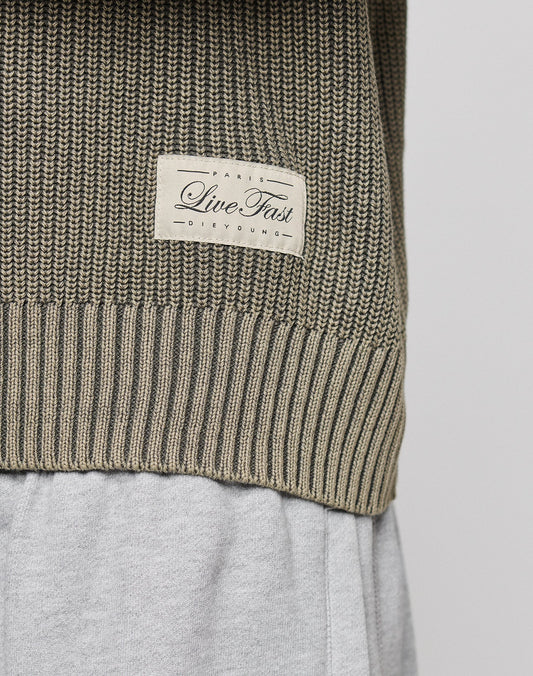 Close-up of the LFDY Knit Sweater by LIVE FAST DIE YOUNG, featuring an oversize fit, dropped shoulders, and a beige patch with "Live Fast" and "Paris DeYoung," styled over light gray pants.