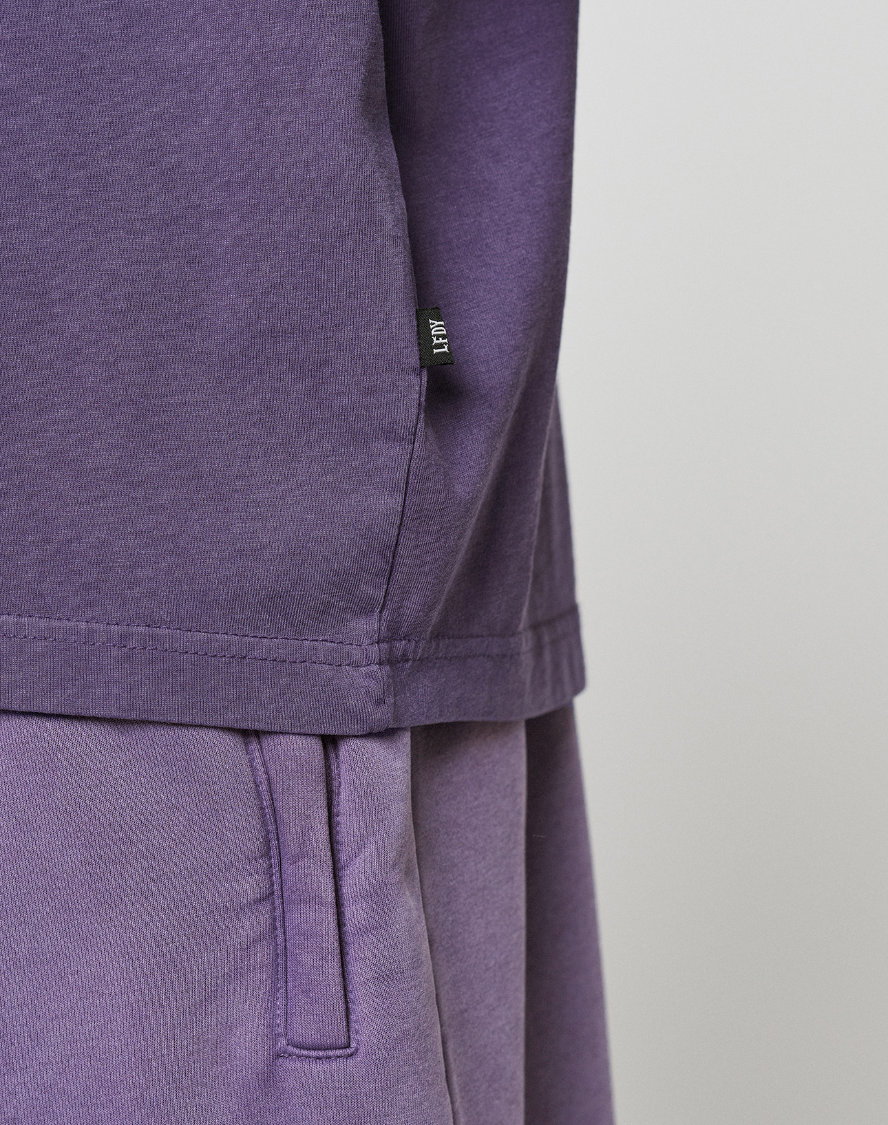 Close-up of a person wearing the LIVE FAST DIE YOUNG Basic Evidence Tee in purple with matching pants, highlighting the side seam, waistband, and small black label. Crafted from 100% Baumwolle for comfort against a plain light background.