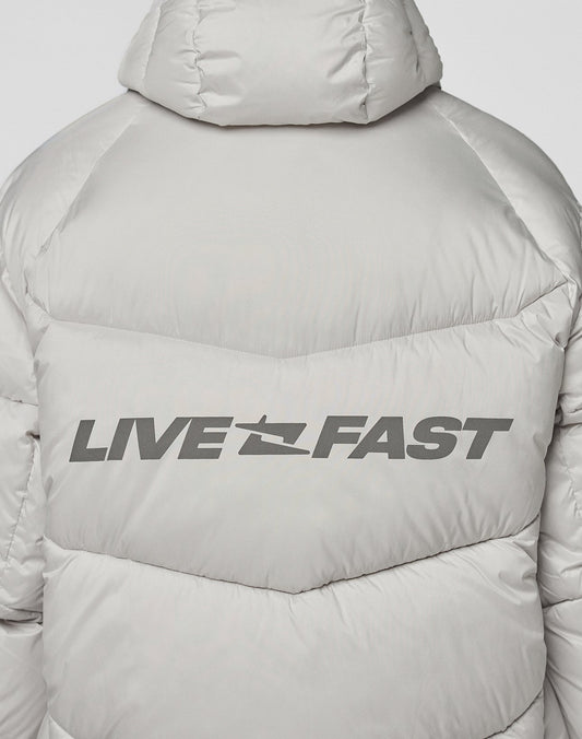 A close-up of the Live Fast Puffer Jacket by LIVE FAST DIE YOUNG shows the light gray, oversized fit with “LIVE FAST” in bold black letters, separated by a jet silhouette and a reflective print on the upper back.