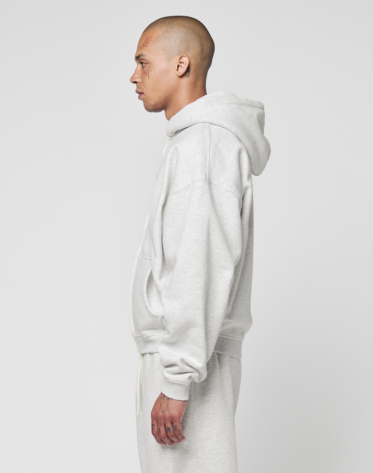 A person with closely shaved hair and facial tattoos stands in profile, wearing the LIVE FAST DIE YOUNG Basic Hooded Zip sweatshirt and matching oversize sweatpants in light gray against a plain white background.