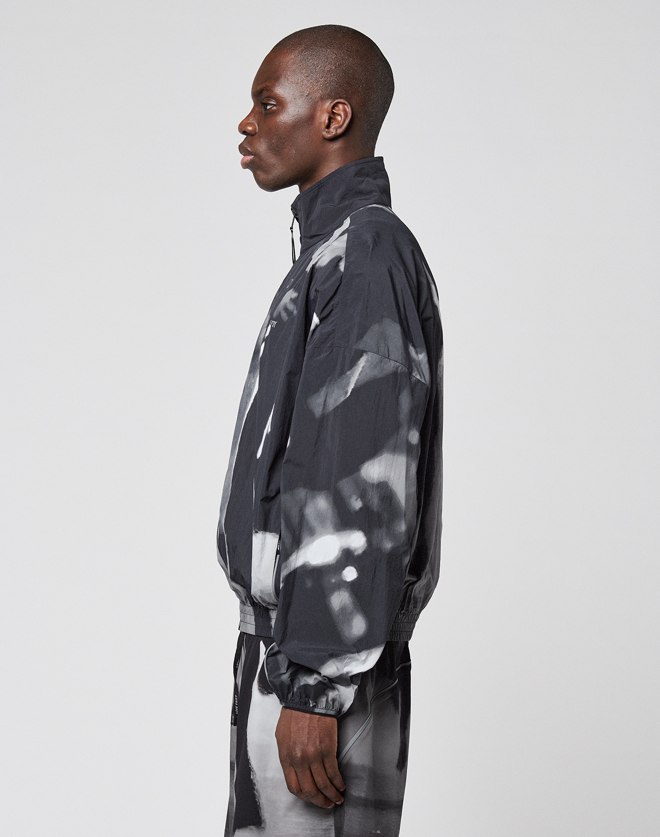 A person stands in profile wearing the LIVE FAST DIE YOUNG LFDY Performance Jacket with matching pants, featuring an allover print and reflective logo, set against a plain light gray background.