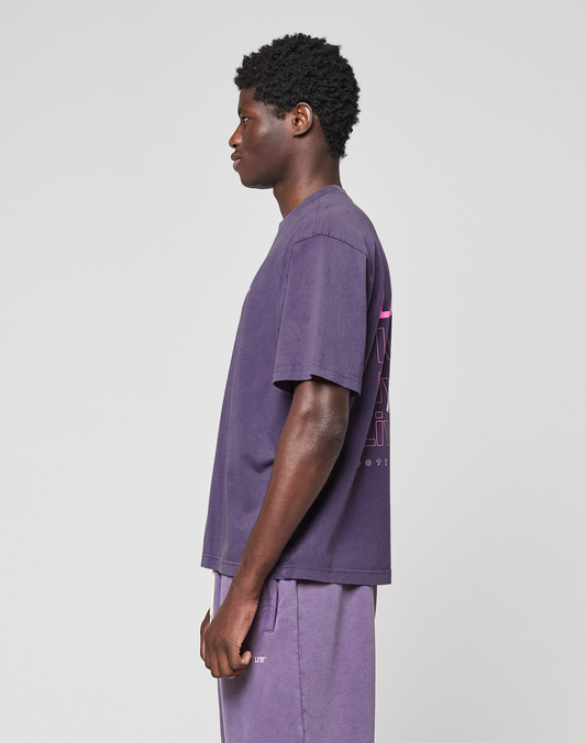 A person in profile against a plain backdrop wears an oversize purple LIVE FAST DIE YOUNG Basic Evidence Tee (100% Baumwolle) and matching shorts; the tee’s back has pink graphic text.