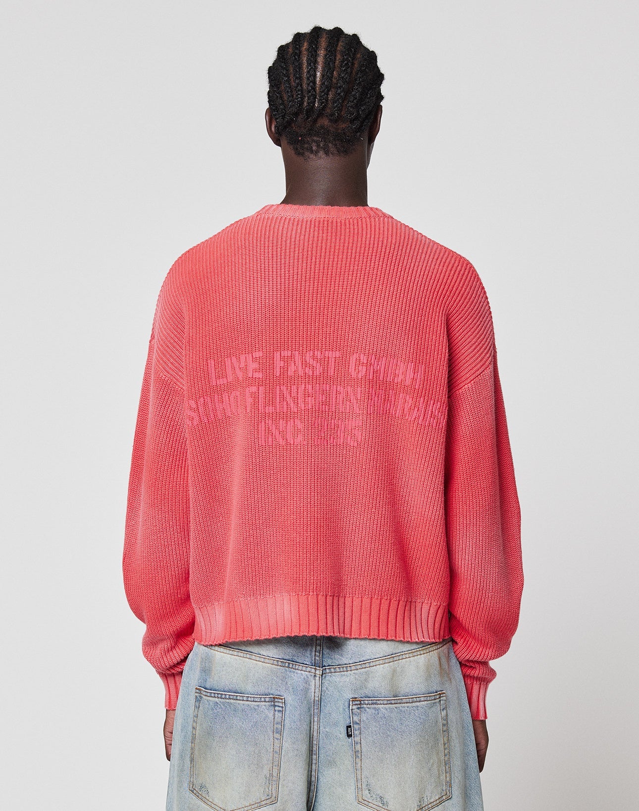 A person with braided hair faces away from the camera, wearing the LFDY Washed Knit Sweater by LIVE FAST DIE YOUNG in a pinkish-red oversize fit with faded text, paired with light blue jeans. Sweater is 100% Baumwolle. Plain light background. | model