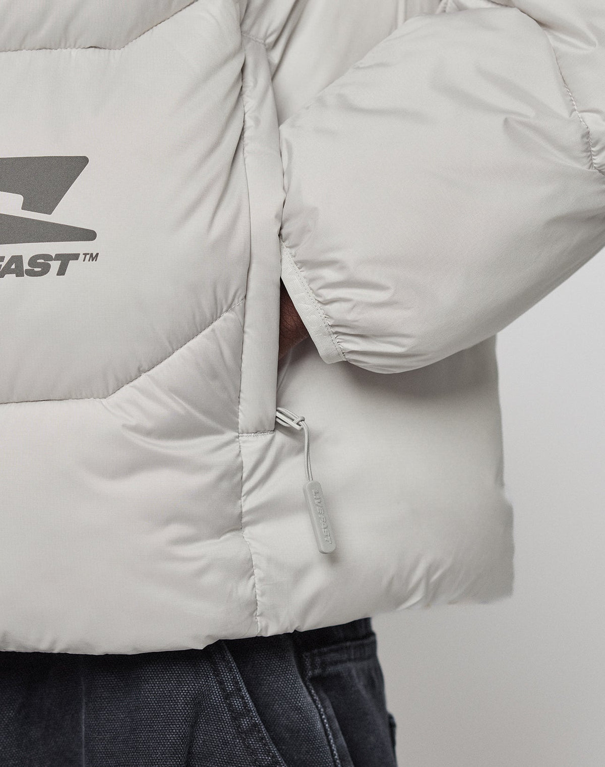 Close-up of a person wearing the LIVE FAST DIE YOUNG Live Fast Puffer Jacket in light grey with visible zipper, chest logo, and hand in a side pocket, paired with dark grey pants.