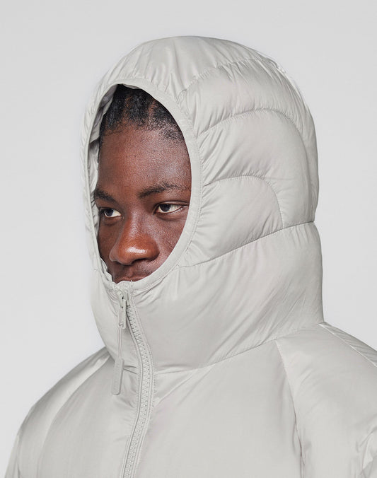 A person wears the LIVE FAST DIE YOUNG Live Fast Puffer Jacket in light gray, featuring an oversized fit and hood zipped up, looking slightly to the side against a plain light background.
