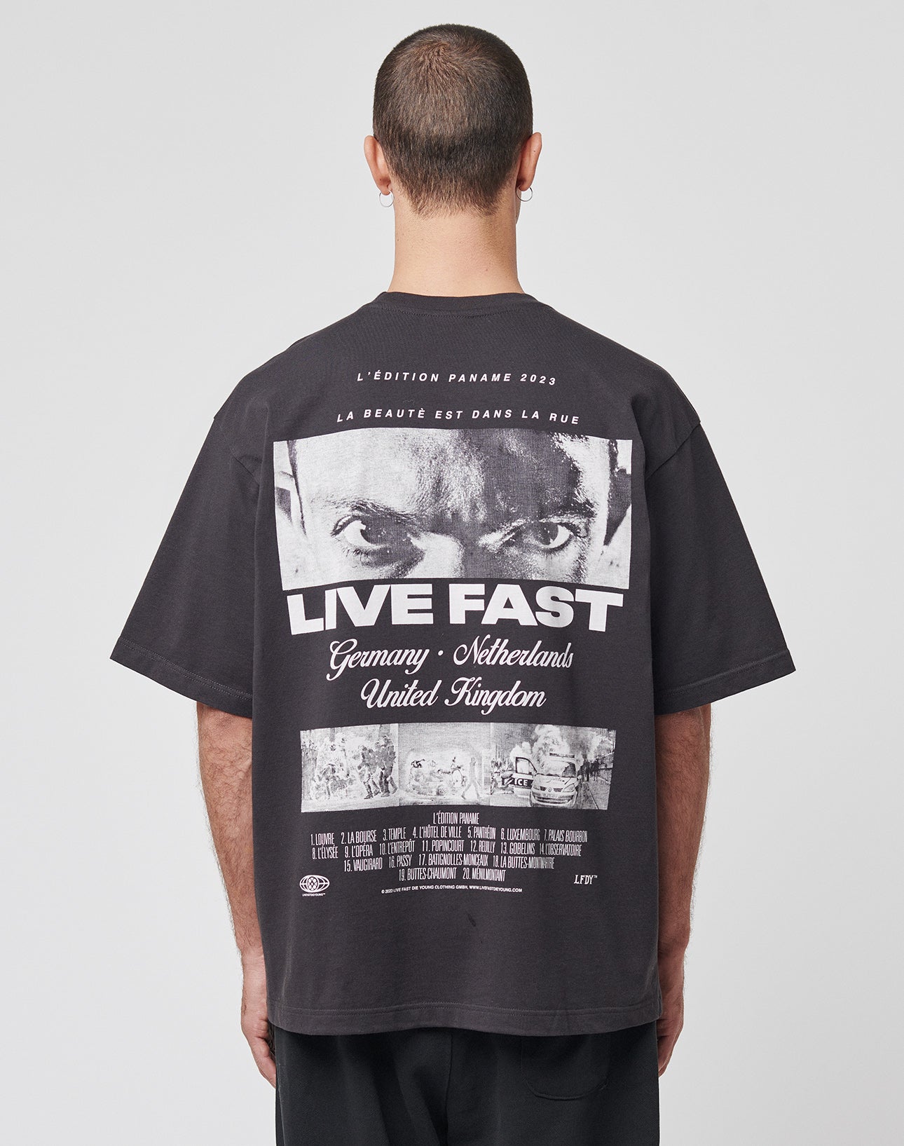 Someone with short hair, facing away, wears the LIVE FAST DIE YOUNG LFDY Cinema Tee—an oversized black Baumwolle t-shirt with a bold backprint of intense eyes, “LIVE FAST,” and the countries Germany, Netherlands, and United Kingdom. | model