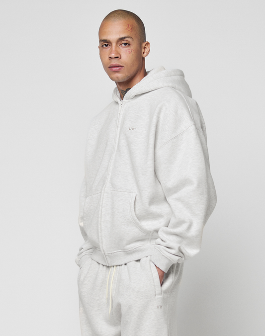 A person wears the LIVE FAST DIE YOUNG Basic Hooded Zip and matching sweatpants, featuring brand logos, standing against a white background with hands in pockets, looking at the camera.