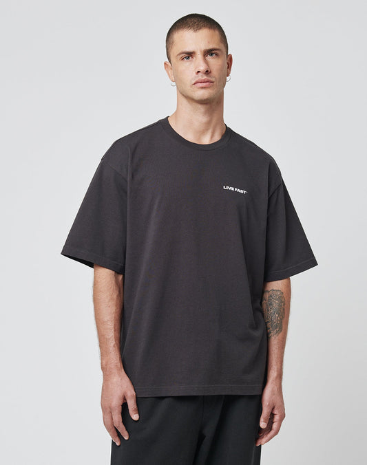 A man with short hair wears the LFDY Cinema Tee by LIVE FAST DIE YOUNG, an oversize black cotton T-shirt featuring a small white LIVIN FAST print on the chest, standing against a plain light gray background and looking to the side.