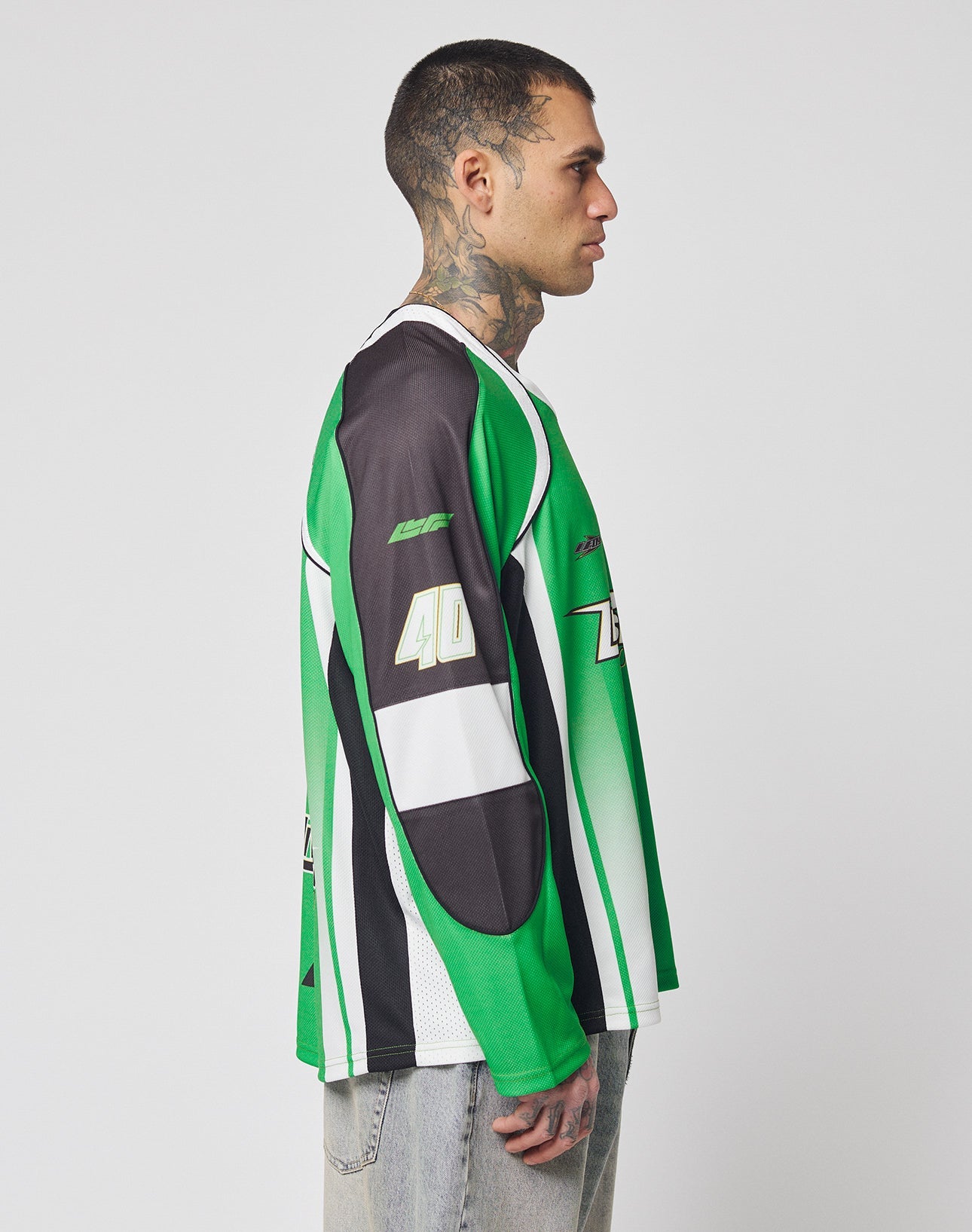 A man with short hair and neck tattoos stands in profile, wearing the LIVE FAST DIE YOUNG LFDY Motorcross Jersey—an oversize green, black, and white long-sleeve with multi-logo prints and 40 on the sleeve—paired with light-washed jeans.