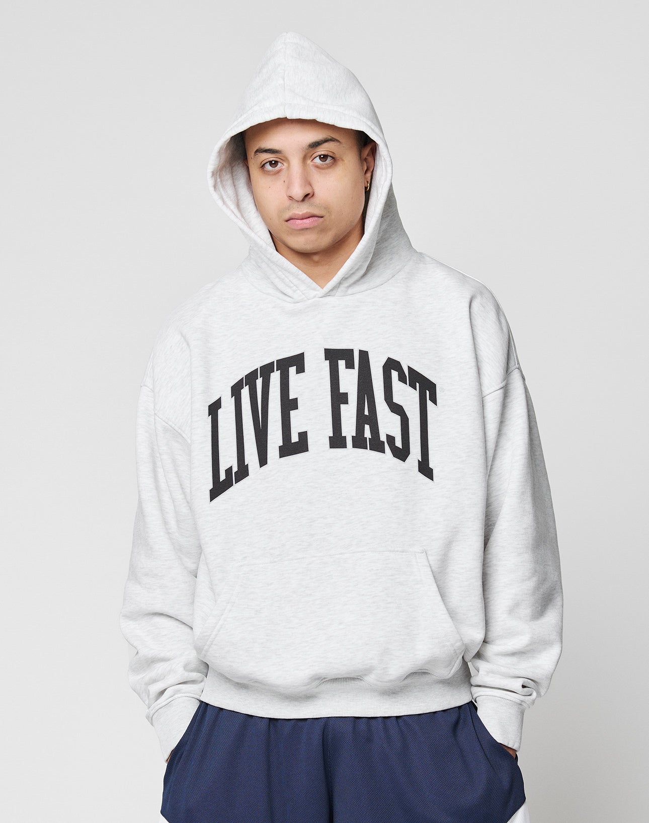 Someone in the LIVE FAST DIE YOUNG Live Fast College Hooded, light gray with bold black LIVE FAST frontprint, stands hands-in-pockets and hood up against a plain light background.