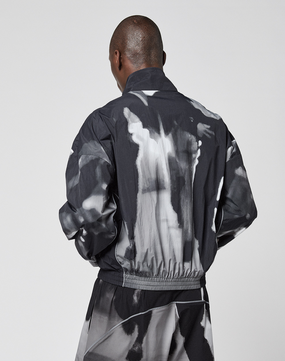 A person with short hair stands with their back to the camera, wearing the black and white LFDY Performance Jacket by LIVE FAST DIE YOUNG, featuring an allover abstract print, against a plain light background.