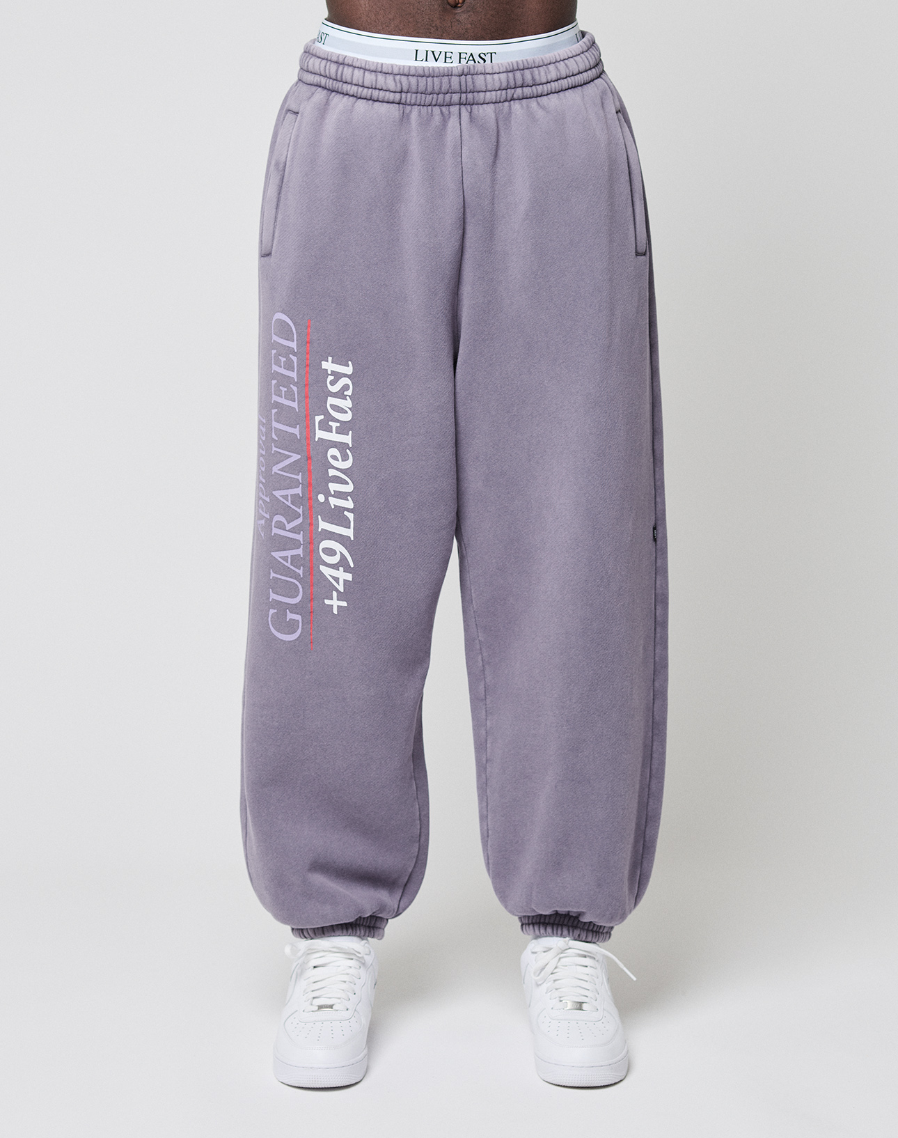 Someone dressed in the LFDY Hotline Loose Jogger by LIVE FAST DIE YOUNG in purple, with “GUARANTEED +49 Live Fast” printed on the leg, pairs them with white sneakers and stands against a plain background.