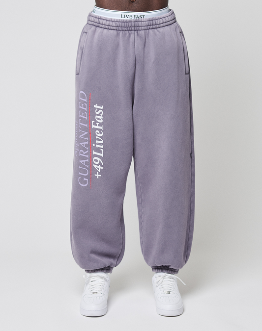 Someone dressed in the LFDY Hotline Loose Jogger by LIVE FAST DIE YOUNG in purple, with “GUARANTEED +49 Live Fast” printed on the leg, pairs them with white sneakers and stands against a plain background.