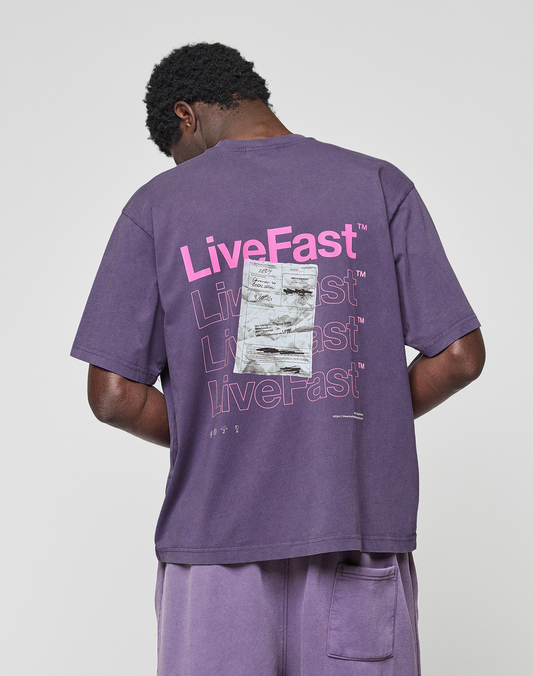 A person faces away, wearing the LIVE FAST DIE YOUNG Basic Evidence Tee in Oversize Fit purple with "LiveFast" printed in pink and white on the back and a silver rectangle patch. Made from 100% Baumwolle.