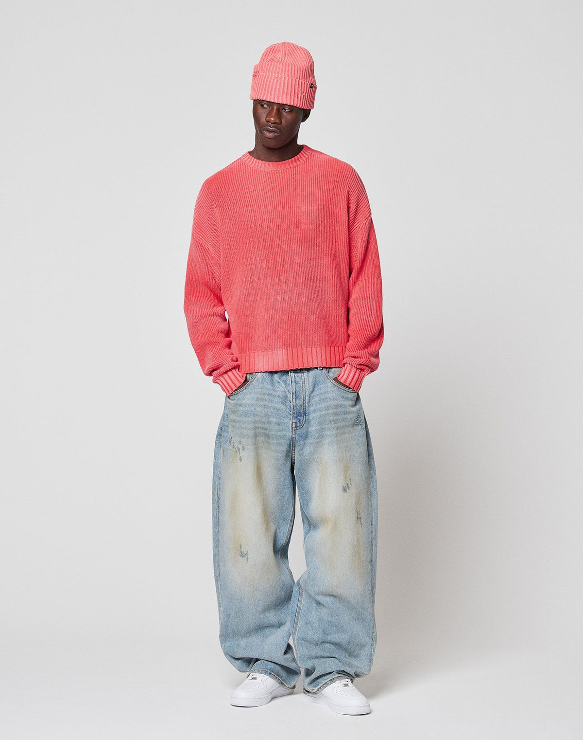 Wearing the LFDY Washed Knit Sweater by LIVE FAST DIE YOUNG, oversized and pink, with very baggy light blue jeans and white sneakers, a person stands against a plain light background, hands in pockets.