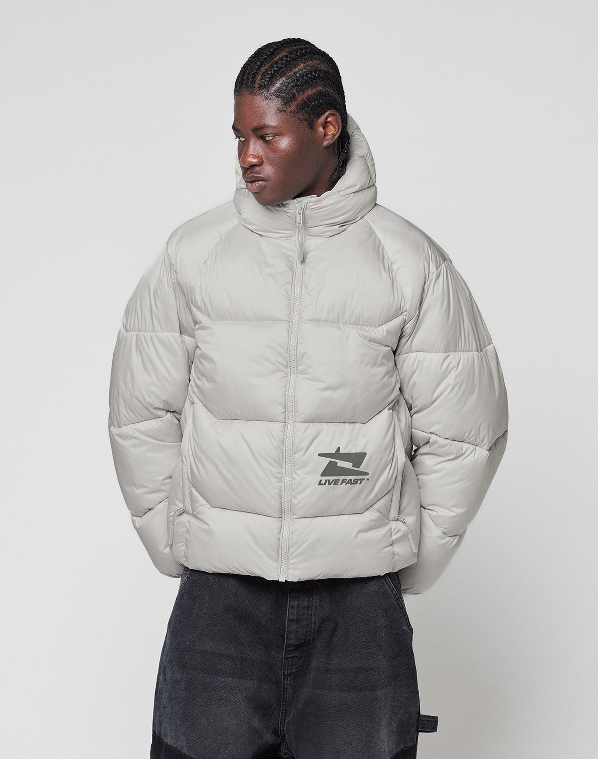 A person with braided hair wears the LIVE FAST DIE YOUNG Live Fast Puffer Jacket in light gray with a high collar and reflective "LIVE FAST" print, paired with dark pants, standing against a plain white background.