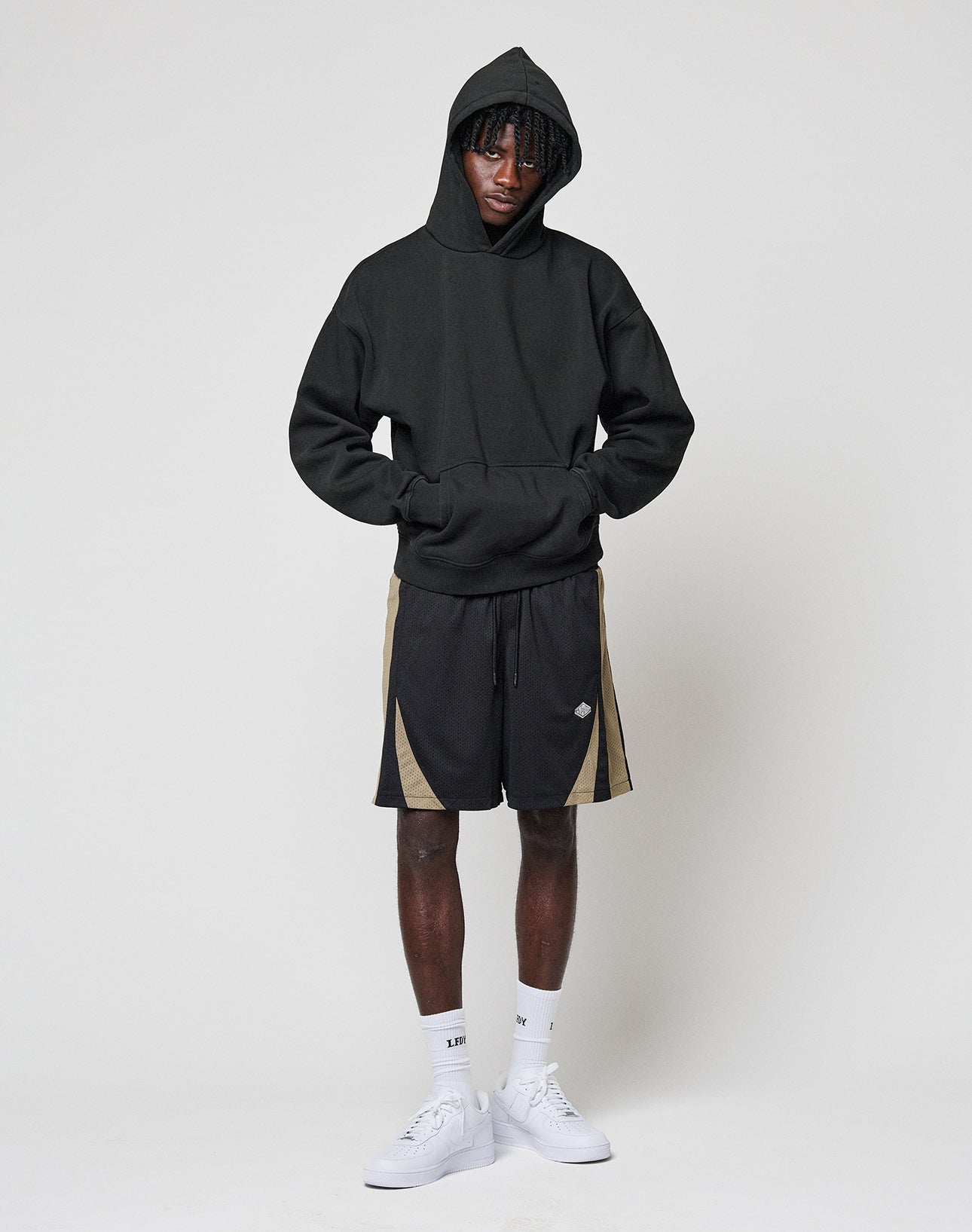 A person stands against a plain light background, hands in the pocket of a LIVE FAST DIE YOUNG Basic Blank Hooded black oversize hoodie, paired with black and beige athletic shorts, white socks, and white sneakers.