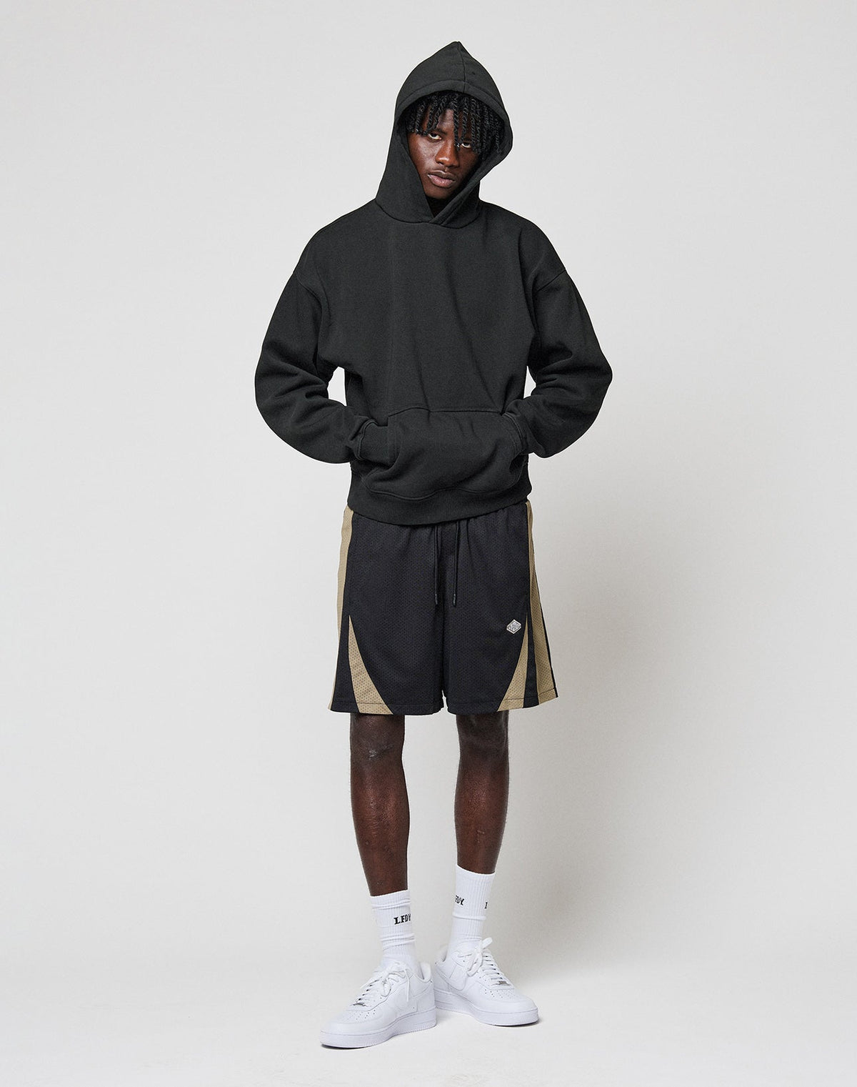 A person stands against a plain light background, hands in the pocket of a LIVE FAST DIE YOUNG Basic Blank Hooded black oversize hoodie, paired with black and beige athletic shorts, white socks, and white sneakers.