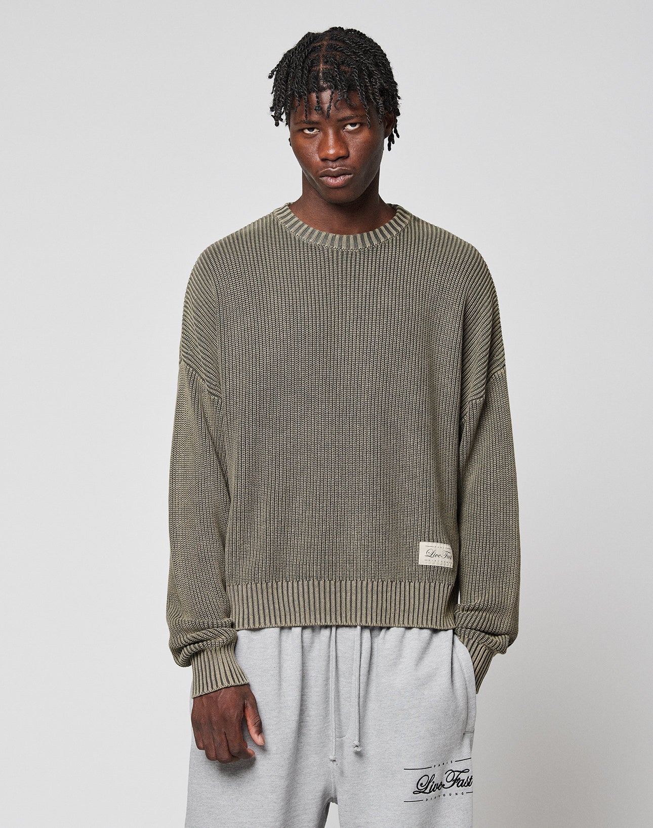 A young man with short dreadlocks wears the LFDY Knit Sweater by LIVE FAST DIE YOUNG—an oversized, ribbed olive-green piece with dropped shoulders—paired with light gray sweatpants against a plain gray background.