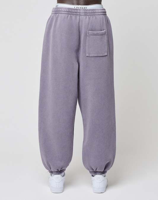 A person wearing the LFDY Hotline Loose Jogger by LIVE FAST DIE YOUNG in lavender with elastic waistband, single patch pocket, leg print, and white sneakers is seen from behind against a plain background.