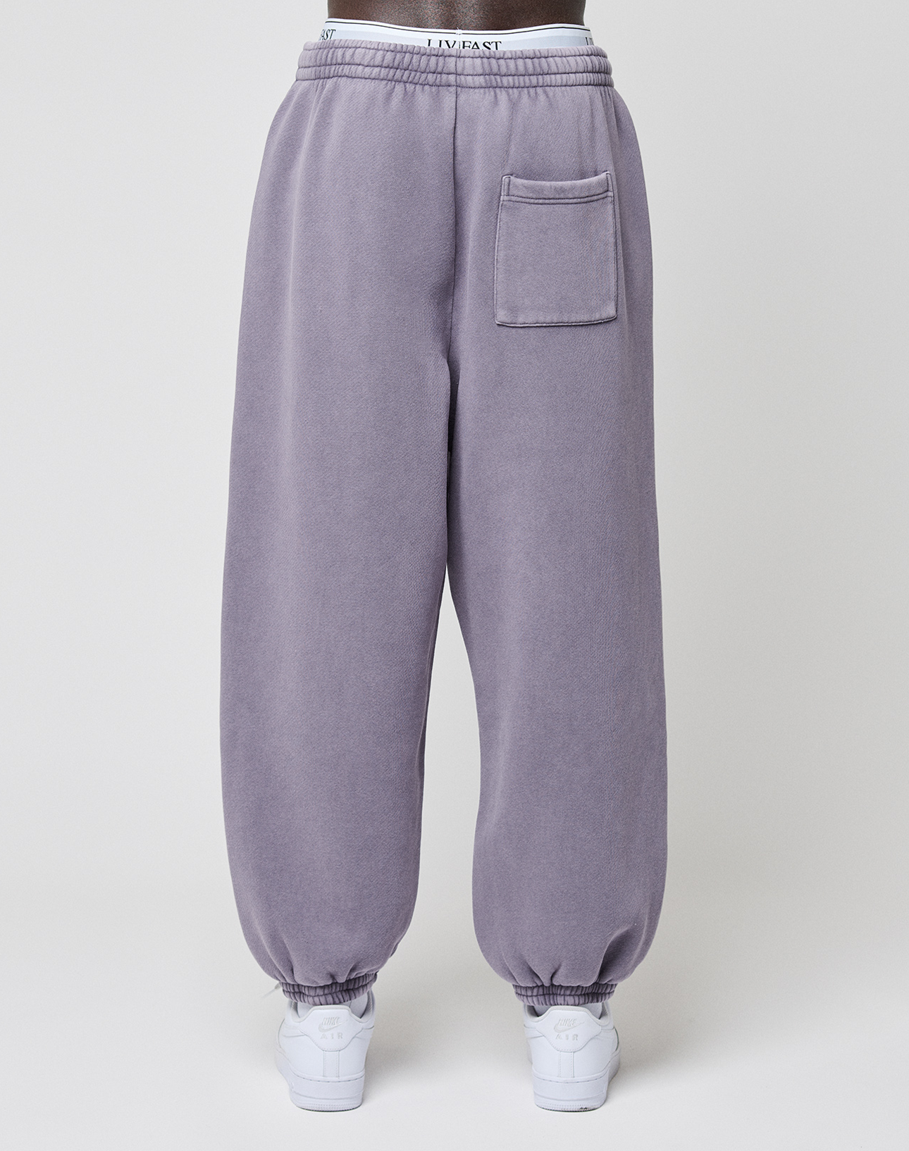 A person wearing the LFDY Hotline Loose Jogger by LIVE FAST DIE YOUNG in lavender with elastic waistband, single patch pocket, leg print, and white sneakers is seen from behind against a plain background.