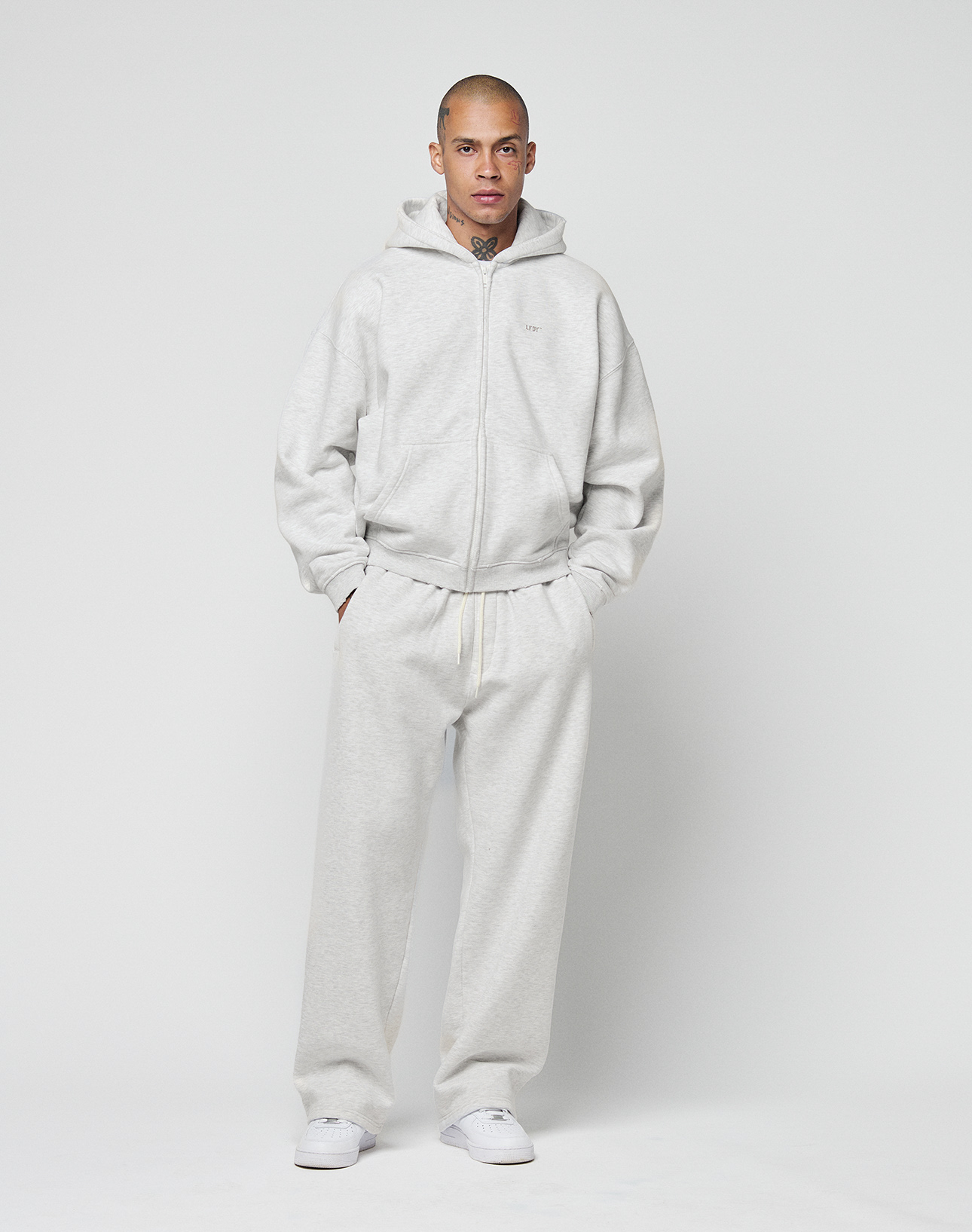 A person with a shaved head stands against a plain white background, hands in pockets, wearing the LIVE FAST DIE YOUNG Basic Hooded Zip in light gray with matching sweatpants and white sneakers.