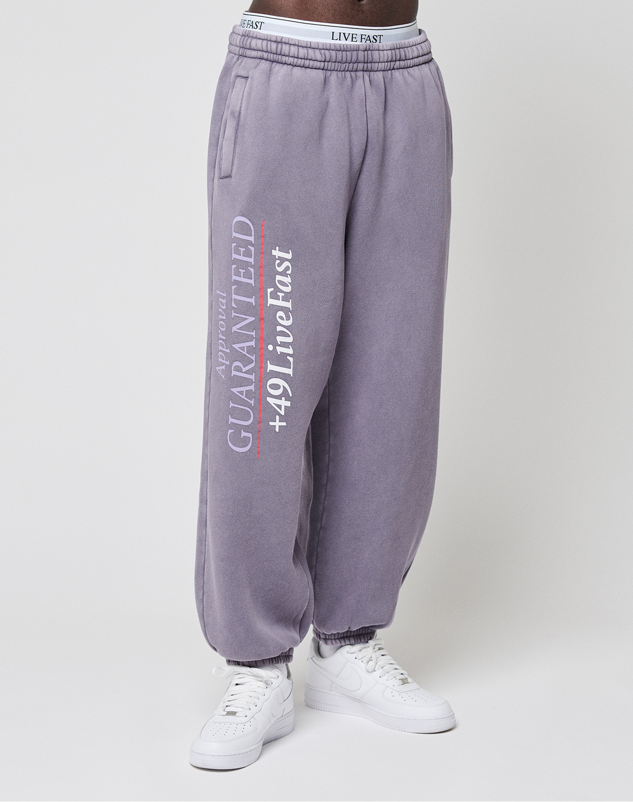 A person wears loose-fitting, purple-grey LFDY Hotline Loose Jogger by LIVE FAST DIE YOUNG with a white and red "Approval GUARANTEED +49 Live Fast" print on the leg, white sneakers, and an Elastikbund for comfort. Only the lower body is shown.