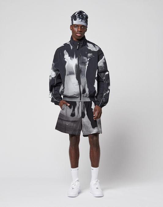 A man stands against a plain background, wearing a black and white LFDY Performance Jacket by LIVE FAST DIE YOUNG with matching shorts, a headband, white socks, and sneakers. He looks at the camera with a relaxed expression.