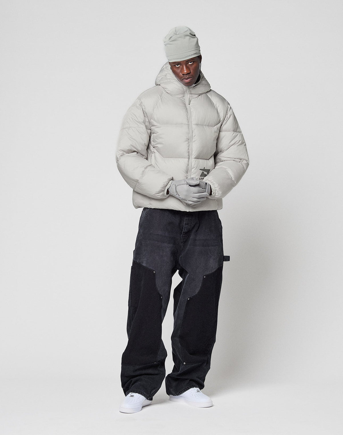 A person wearing the LIVE FAST DIE YOUNG Live Fast Puffer Jacket in light gray, with matching beanie and gloves, loose black pants, and white sneakers stands against a plain white background.
