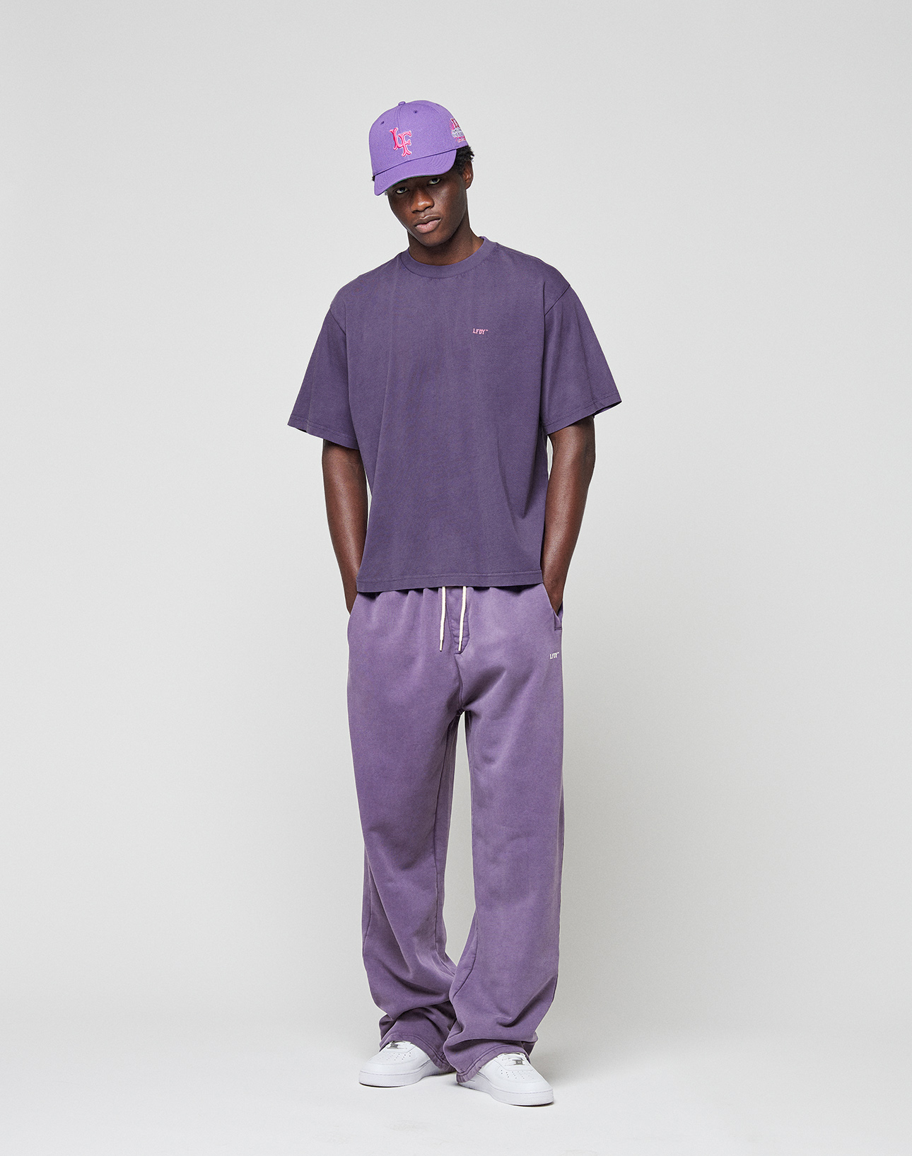 A person faces forward with hands in pockets, wearing the LIVE FAST DIE YOUNG Basic Evidence Tee in an oversize fit and matching sweatpants, paired with a purple baseball cap and white sneakers against a plain background.