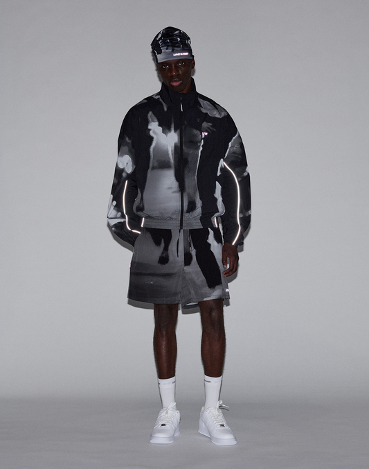 A man stands against a plain background wearing LIVE FAST DIE YOUNG’s LFDY Performance Shorts and a matching Regular Fit black and white abstract-patterned jacket with a reflective logo print, white socks, sneakers, and a headband for comfort.
