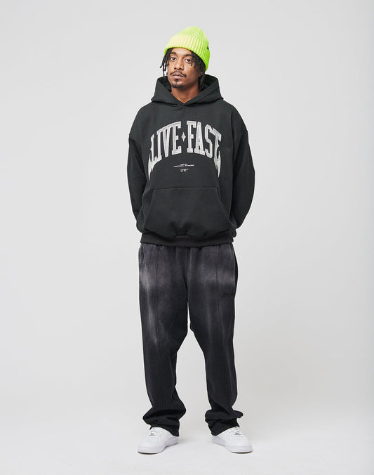 A person stands against a plain background wearing a neon green oversize beanie, the LIVE FAST DIE YOUNG Live Fast Axe Hooded black hoodie with LIVE FAST on it, loose black Baumwolle pants, and white sneakers. Their hands are in their pockets and they look to the side.