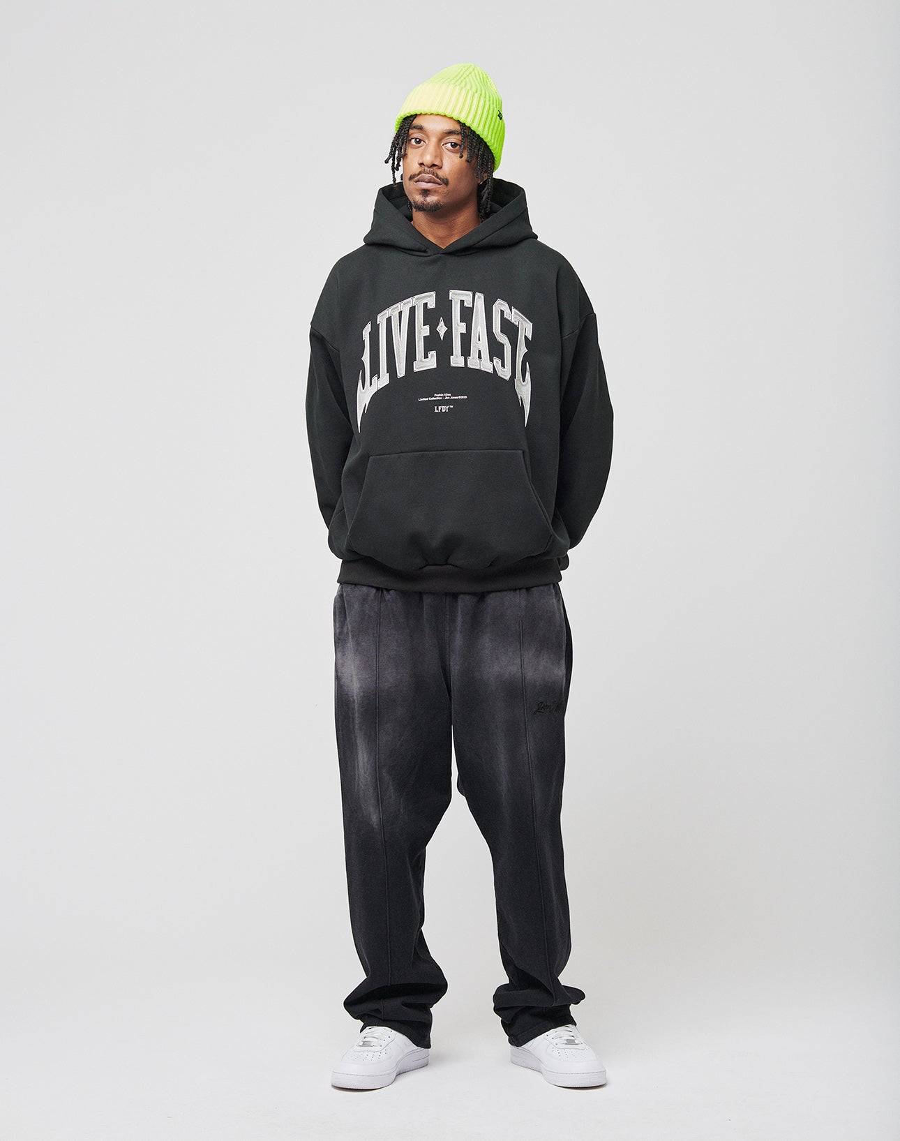 A person stands against a plain background wearing a neon green oversize beanie, the LIVE FAST DIE YOUNG Live Fast Axe Hooded black hoodie with LIVE FAST on it, loose black Baumwolle pants, and white sneakers. Their hands are in their pockets and they look to the side.