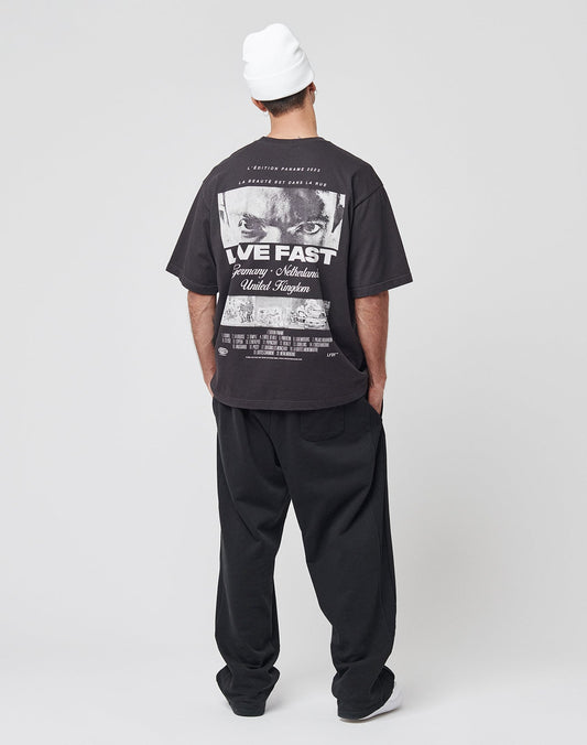 A person in a white beanie, loose black pants, and the LIVE FAST DIE YOUNG LFDY Cinema Tee with a back graphic stands facing away from the camera against a plain light grey background.