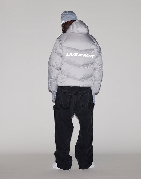 A person faces away, wearing the LIVE FAST DIE YOUNG Live Fast Puffer Jacket in oversize light gray with reflective "LIVE FAST" print on the back, black jeans, white shoes, and a matching beanie, set against a plain light background.