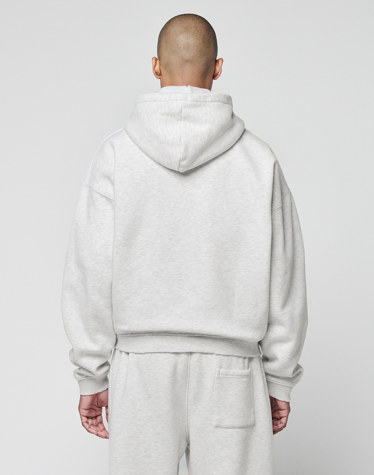 A person with a shaved head stands facing away from the camera, wearing the LIVE FAST DIE YOUNG Basic Hooded Zip in light gray with matching sweatpants, featuring subtle LFDY branding. The background is plain white.