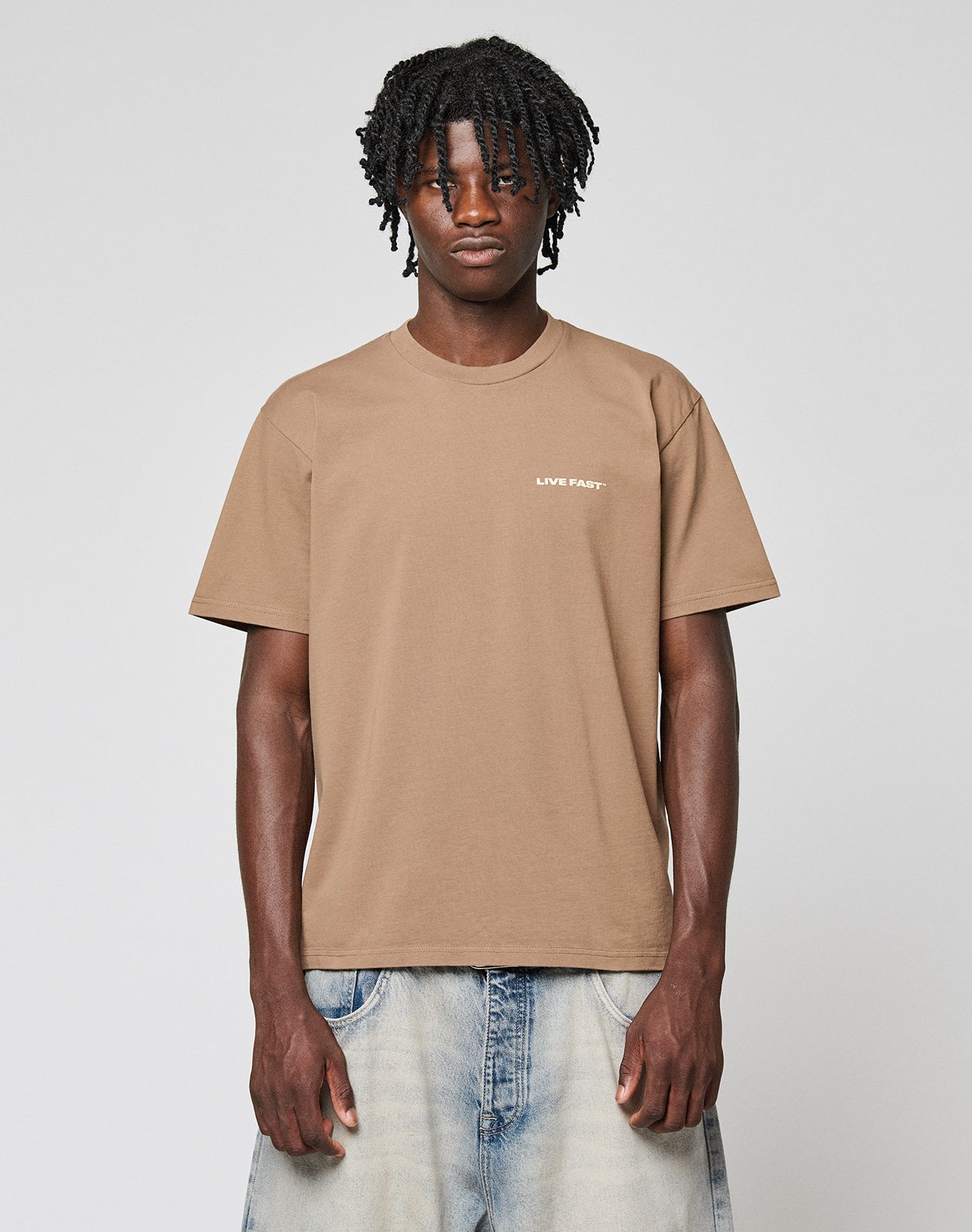A man with short dreadlocks wears the LIVE FAST DIE YOUNG Live Fast Plain Tee, a tan regular fit T-shirt with front print, made from 100% Baumwolle, paired with light blue baggy jeans against a plain light gray background.