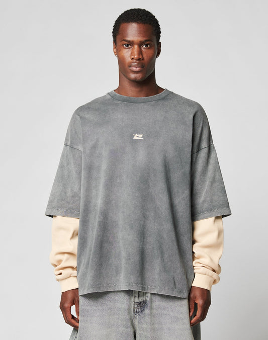 A man stands against a plain background wearing the LIVE FAST DIE YOUNG LFDY Double Layered Longsleeve in an oversize fit with a small white chest logo, paired with gray pants.