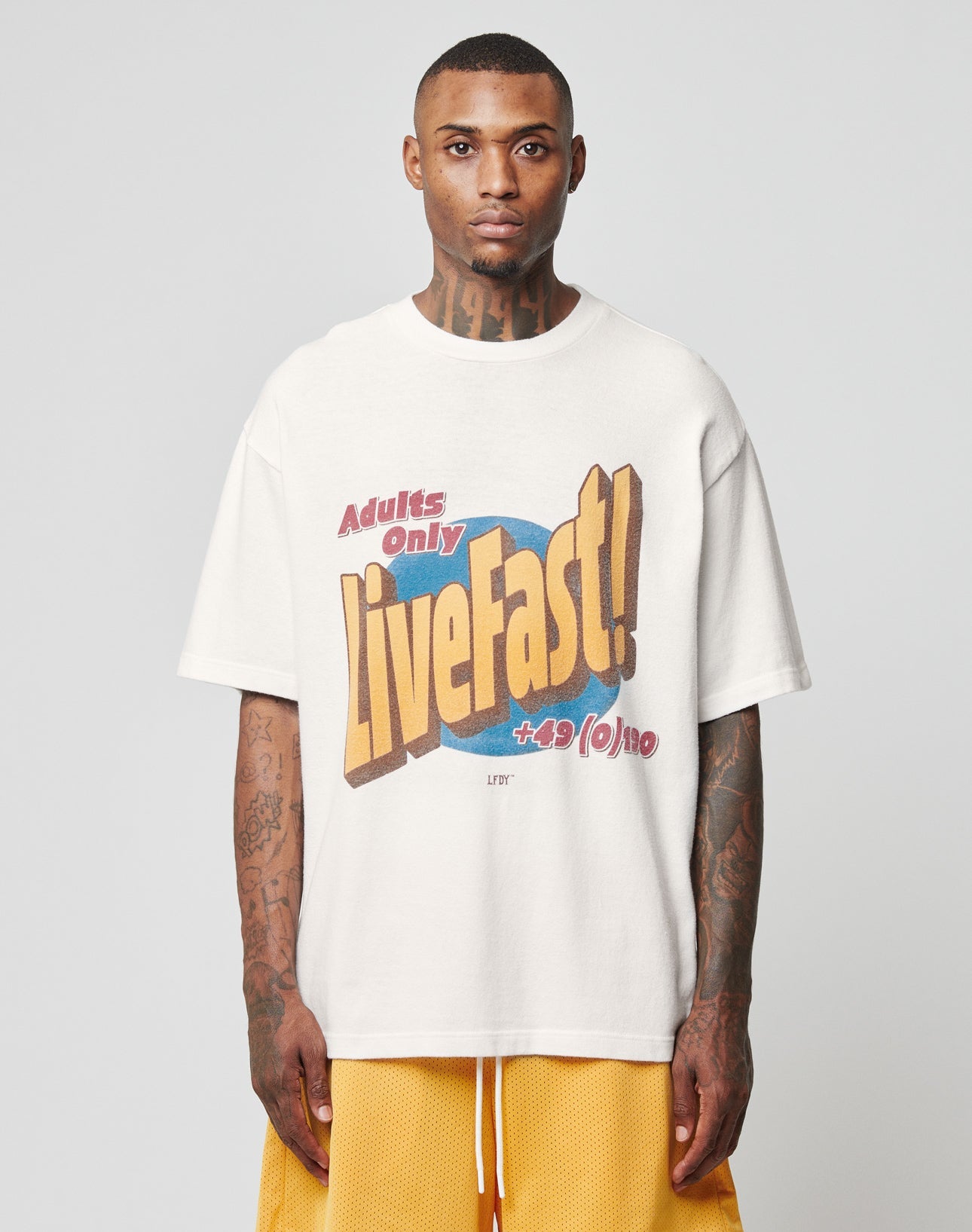 A person faces forward in a loose white LFDY Adults Only Tee by LIVE FAST DIE YOUNG, made of 100% cotton with “Adults Only Live Fast! +9/(o)no” in bold colors, paired with loose yellow shorts. The background is plain light gray.