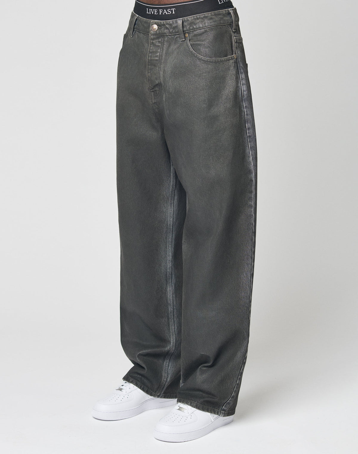 A person in LIVE FAST DIE YOUNG’s LFDY Coated Baggy Denim—baggy, wide-leg, dark grey 100% Baumwolle jeans—paired with white sneakers and LIVE FAST waistband black underwear stands against a plain white background. | model