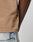 Close-up of the Live Fast Plain Tee by LIVE FAST DIE YOUNG, a beige 100% cotton T-shirt with a small black tag on the seam, paired with light blue denim jeans. Only the lower shirt, part of an arm, and jeans are visible.